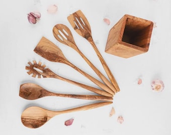 Puede incluir: Un conjunto de utensilios de cocina de madera, que incluyen una espátula ranurada, una cuchara y un tenedor para pasta, dispuestos sobre una superficie blanca. También hay un soporte de madera para utensilios y dientes de ajo.