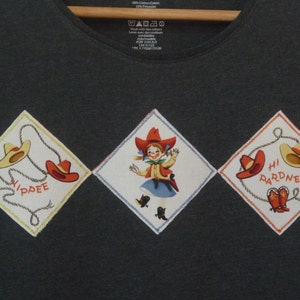 May include: A dark gray t-shirt with three diamond-shaped patches. The patches feature a cowboy hat, a girl in a cowboy outfit, and a cowboy hat with the text "Hi Pardner".