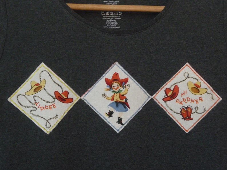 May include: A dark gray t-shirt with three diamond-shaped patches. The patches feature a cowboy hat, a girl in a cowboy outfit, and a cowboy hat with the text "Hi Pardner".
