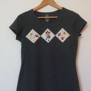May include: A dark gray t-shirt with three embroidered patches. The patches are diamond-shaped and feature a cowboy, a lasso, and the words "Mi Vaquero".