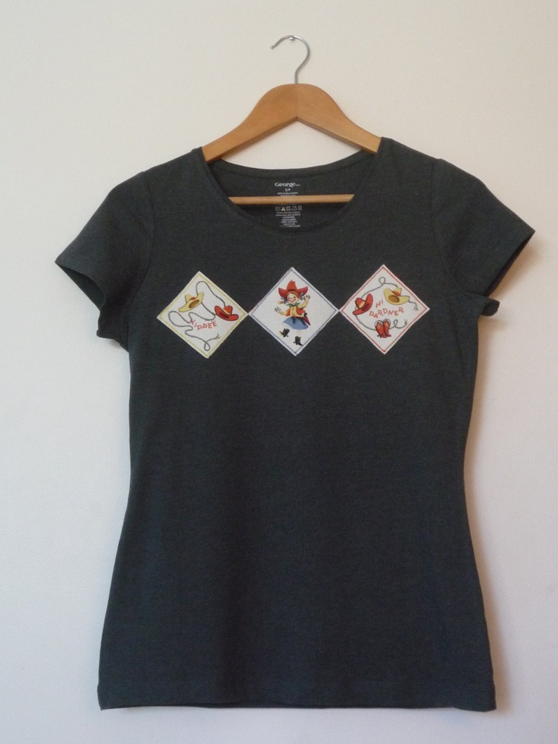 May include: A dark gray t-shirt with three embroidered patches. The patches are diamond-shaped and feature a cowboy, a lasso, and the words "Mi Vaquero".
