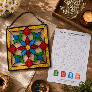 Puede incluir: Un panel de vidriera cuadrado con un círculo central amarillo rodeado de pétalos rojos, azules y verdes dentro de un borde amarillo, suspendido por una cadena negra. Al lado hay un patrón de papel titulado Ruby Blossom Stained Glass Pattern.