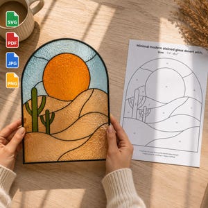 Op de afbeelding: Een boogvormig glas-in-loodpaneel met een woestijnlandschap, een feloranje zon, beige zandduinen en twee groene cactussen. Daarnaast ligt een geprint patroonblad met het opschrift Minimal modern stained glass desert arch.