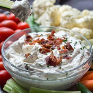 May include: A clear glass bowl filled with a creamy white dip topped with crispy bacon bits and chopped green onions. The dip is surrounded by various vegetables, including celery, carrots, and cauliflower.