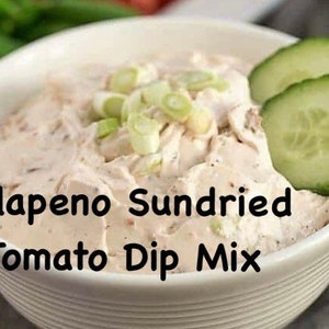 May include: A white bowl filled with a creamy dip, topped with green onions and cucumber slices. The bowl has a text label that reads "Jalapeno Sundried Tomato Dip Mix".