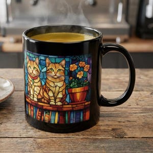 Pode incluir: Caneca de café de cerâmica preta com um design de vitral com dois gatos laranja e flores laranja. A caneca contém uma bebida quente, e vapor sobe do topo. Um croissant está em um prato.