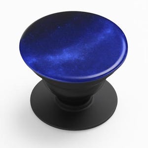 May include: A black phone grip with a round, dark blue top featuring a galaxy design. The grip has a collapsible feature and a circular base. The galaxy design has shades of blue and white, creating a celestial aesthetic.