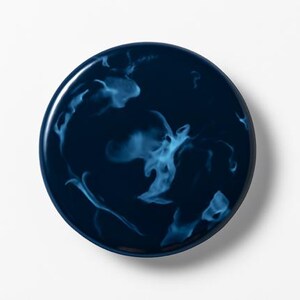 Peut inclure: Une assiette décorative ronde bleu foncé avec une finition brillante. Elle présente un motif tourbillonnant et marbré en bleu clair et blanc, créant un design unique. L'assiette est posée sur un fond blanc uni. Diamètre d'environ 20 cm.
