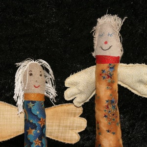 Può includere: Due angeli di stoffa fatti a mano con le ali. Un angelo indossa un tessuto blu e oro con stelle. L'altro angelo indossa un tessuto marrone con stelle rosse e dorate.