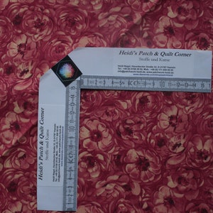 Può includere: Un primo piano di un tessuto rosso con un motivo floreale. Il tessuto è un cotone per trapunte con un motivo ripetitivo di rose rosse e gialle. Un righello è presente nell'immagine per mostrare la scala del tessuto. Il testo sul righello recita "Heidi's Patch & Quilt Corner" e "Stoffe und Kurse".