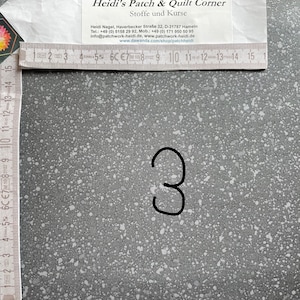 May include: A close-up of a grey fabric with white speckles. The fabric is marked with the number "3" in black marker.