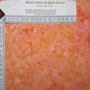 Op de afbeelding: Een close-up van een stof met een oranje en roze batikpatroon. De stof wordt gemeten met een liniaal, wat een lengte van 38 cm laat zien.