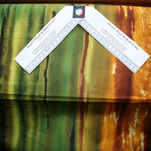 Può includere: Un tessuto con un design simile all'acquerello che presenta strisce verticali in tonalità di verde, giallo, arancione e marrone. Il tessuto è piegato a metà e un righello è posizionato sopra. Il righello è etichettato "Heidi's Patch & Quilt Corner".