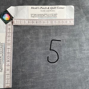 Könnte beinhalten: Nahaufnahme eines grauen Stoffes mit der Zahl 5 in schwarzem Stift geschrieben. Im Hintergrund ist ein Lineal mit dem Text "Heidi's Patch & Quilt Corner" und Kontaktinformationen zu sehen.
