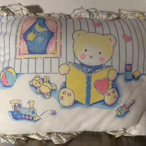 May include: A rectangular pillow with a whimsical design featuring a yellow teddy bear reading a book. The pillow has a blue and white striped background with a window scene, a toy train, and a ball. The edges are ruffled.