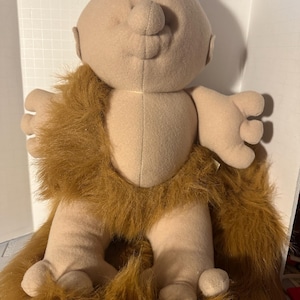 May include: A soft toy doll with a tan body, a large nose, and white hair. The doll has a brown fur-like chest and leg covering. The doll's arms and legs are short and stubby. The doll is a whimsical, handmade item.