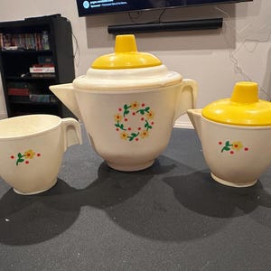 Op de afbeelding: Een vintage crèmekleurige theeset met gele accenten. De set bevat een theepot met een gele deksel, een melkkan en een suikerpot. Elk stuk is versierd met een bloemmotief. De theepot heeft een bloemenkrans.