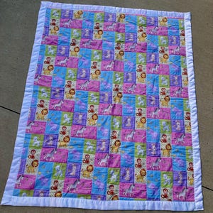 May include: A rectangular patchwork quilt featuring a repeating pattern of cartoon animals including lions, zebras, and monkeys. The quilt is composed of alternating pink, blue, and green squares with a solid white fabric border around the edge.