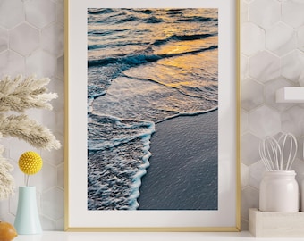 May include: A framed art print featuring a coastal scene. The image captures the ocean waves meeting the shore, with golden sunlight reflecting on the water. The frame is gold, and the overall aesthetic is serene and calming.