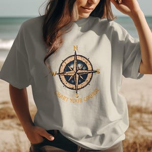 May include: A light gray t-shirt featuring a vintage compass design with the text "Chart Your Course" in a gold color. The compass has a blue center and gold accents. The shirt is worn by a person on a beach.