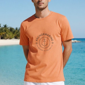 Puede incluir: Camiseta naranja de manga corta con un gráfico circular que dice "ANCHORMARINE" alrededor de un símbolo de ancla. El texto "ESCAPE TO THE SEA" está debajo. La camiseta está hecha de un material suave.