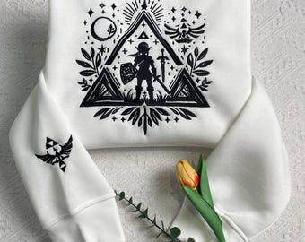 May include: White sweatshirt featuring a black graphic design of a video game character with a sword and shield, set within a triangle and other geometric shapes. The sleeve has a matching emblem. A yellow and orange tulip and greenery are in the foreground.