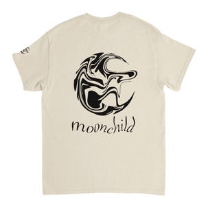 Könnte beinhalten: Cremefarbenes T-Shirt mit einem schwarz-weißen abstrakten Design und dem Wort "moonchild" in Schreibschrift. Das Design befindet sich auf der Rückseite des Shirts, mit einer kleinen Grafik auf dem linken Ärmel.