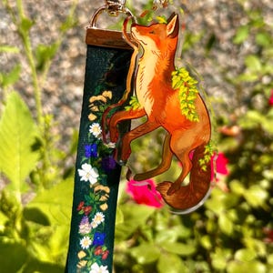 Puede incluir: Un llavero con una correa de tela verde oscuro decorada con un patrón floral colorido y un dije de acrílico transparente con forma de zorro adornado con hojas verdes. El zorro es naranja con detalles blancos y tiene un cierre de metal dorado.