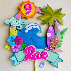 Peut inclure: Un cake topper coloré sur le thème tropical. Il présente une vague bleue, un soleil avec le chiffre 9, le mot "Aloha" et le nom "Rae". Comprend un palmier, des planches de surf et des fleurs d'hibiscus.