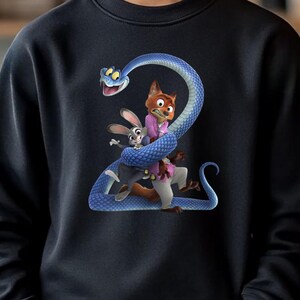 May include: Black sweatshirt featuring a cartoon graphic of a blue snake with a fox and a rabbit. The sweatshirt is worn by a person with light brown hair, smiling in front of a background of plants.