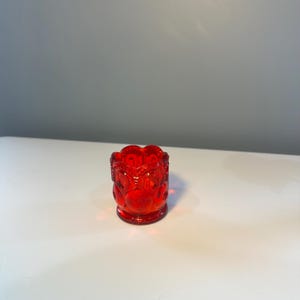 May include: A small, ruby-red glass votive candle holder. The candle holder has a detailed, embossed design with a scalloped top edge. The glass is transparent, allowing light to pass through.