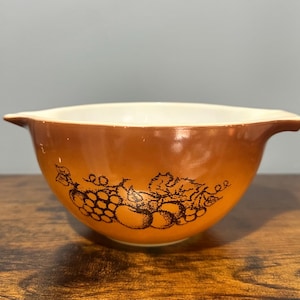 Puede incluir: Un cuenco mezclador Pyrex marrón vintage con interior blanco. El cuenco presenta un exterior degradado marrón con un diseño de uvas y hojas negras. El cuenco tiene dos asas.