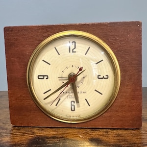 Puede incluir: Un reloj vintage General Electric Telechron con marco de madera. La esfera del reloj es de color crema con números y manecillas negras. El reloj tiene un bisel dorado y las palabras "General Electric Telechron" están impresas en la esfera.