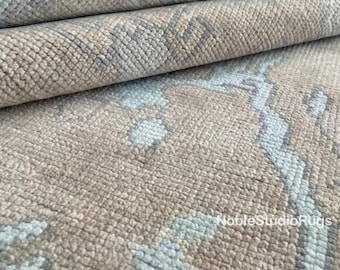 May include: Close-up of a beige and light blue patterned rug. The rug has a textured surface with a repeating pattern of light blue and beige shapes. The rug is folded over in layers, showing the texture and design.