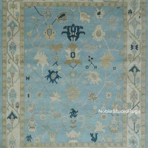 May include: A rectangular, light blue rug with an intricate floral and geometric pattern. The rug features a central field of light blue, bordered by a cream-colored frame with floral motifs. The design includes navy blue, beige, and cream-colored floral elements.