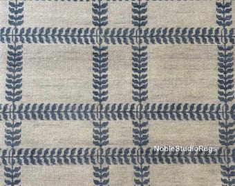 May include: A beige rug with a repeating pattern of blue leaf-like designs. The pattern forms a grid-like structure. The rug has a textured appearance. The text "Noble StudioRugs" is visible on the rug.