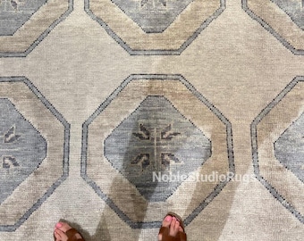 May include: A beige and blue patterned rug with geometric designs. The rug features octagonal shapes in beige and blue, with a central floral motif. The rug's border is also blue. The image includes a person's feet wearing brown sandals and blue jeans.