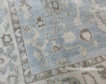 May include: A close-up of a light blue rug with a detailed pattern. The rug features a central field of light blue with a border of gray and beige. The design includes floral and geometric elements. The text "NobleStudioRugs" is visible.