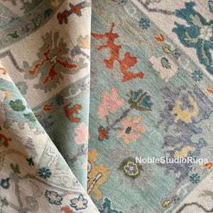 Peut inclure: Gros plan sur un tapis plié à motifs floraux. Le tapis présente un fond bleu clair avec une bordure crème et des accents orange, rose et sarcelle. Le texte "NobleStudioRugs" est visible.