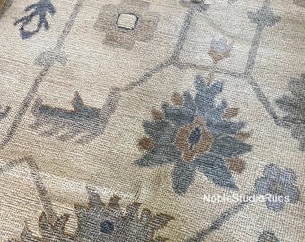 May include: Close-up of a beige rug with a detailed floral pattern in shades of blue, brown, and gray. The rug has a textured appearance and features a repeating design of stylized flowers and geometric shapes. The rug is a decorative home decor item.