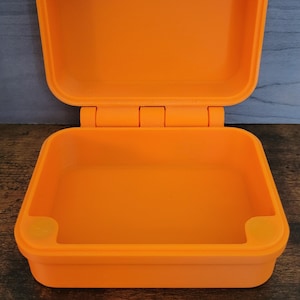 May include: Bright orange, open, hinged container. The rectangular box has rounded corners and a matching lid. The lid is open, revealing the interior. The container is made of a solid, opaque material. The box is sitting on a wooden surface.
