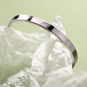 Può includere: Un braccialetto a cerchio minimalista color argento. Il braccialetto ha un profilo piatto e rettangolare ed è esposto su un letto di carta bianca stropicciata su uno sfondo verde chiaro. Il design semplice del braccialetto suggerisce un'estetica moderna.