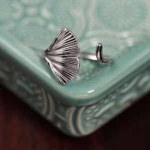 Puede incluir: Anillo plateado con diseño de hoja de ginkgo. Se encuentra sobre una fuente de cerámica verde azulado claro con un patrón decorativo. La hoja de ginkgo está detallada con finas líneas.