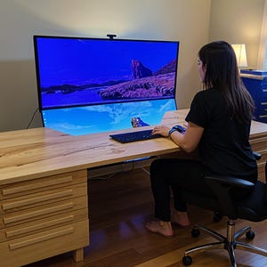 Op de afbeelding: Een lichtgekleurde houten bureau met meerdere laden en een grote computermonitor met een landschap. Een persoon zit aan het bureau en gebruikt een toetsenbord. Het bureau staat in een kamer met een raam en een blauwe kast.