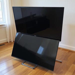 May include: Two large, black flat-screen televisions, one stacked on top of the other. The bottom TV is angled, supported by a silver metal stand. The screens reflect the surrounding room, set on a hardwood floor.