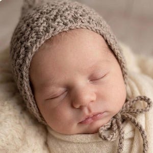May include: A light brown, textured, hand-crocheted bonnet with a pointed top and matching braided ties that fasten under the chin. The bonnet features a dense, woven stitch pattern and covers the head, framing the face with a simple, clean edge.