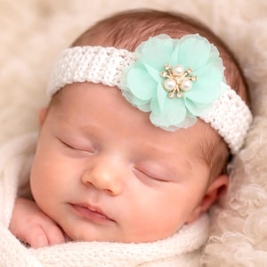 Op de afbeelding: Een slapende baby met een witte gehaakte hoofdband met een grote, lichtgroene chiffon bloem. De bloem is versierd met imitatieparels en strass-steentjes. De baby is gewikkeld in een zachte, witte deken.