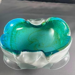 May include: A glass bowl featuring a vibrant turquoise interior with swirling gold-toned flecks and a thick, opaque white exterior. The bowl has a distinct four-lobed, scalloped rim design with a smooth, polished finish and a heavy, rounded base.