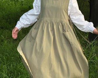 May include: An olive green apron dress with two front pockets and tie closures. The dress is layered over a white long-sleeved blouse. The dress has a gathered skirt and a scoop neckline. The image is taken outdoors in a grassy area.