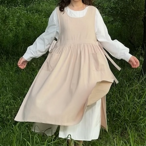 Könnte beinhalten: Ein hellbeige Schürzenkleid mit zwei Vordertaschen und seitlichen Bindebändern. Das Kleid ist über einer weißen Langarmbluse und einer weißen Hose geschichtet. Das Outfit wird im Freien mit grünem Laub im Hintergrund getragen.